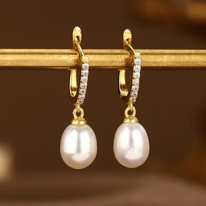 May include: A pair of gold hoop earrings with a single pearl drop on each earring. The hoops are adorned with small clear stones.