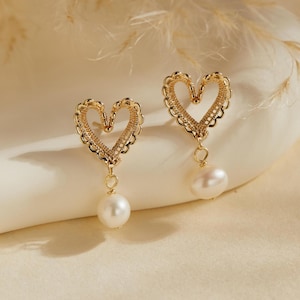 Può includere: Un paio di orecchini a forma di cuore color oro con un delicato design traforato. Ogni orecchino presenta una perla bianca a goccia pendente. Gli orecchini sono esposti su una superficie color crema.