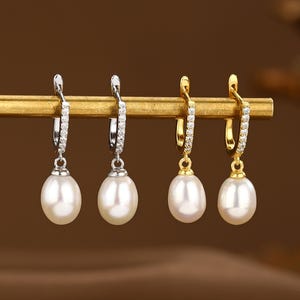 May include: Four pairs of pearl drop earrings with a diamond encrusted hoop. Two pairs of earrings have a gold hoop and two pairs of earrings have a silver hoop. The earrings are displayed on a gold bar.