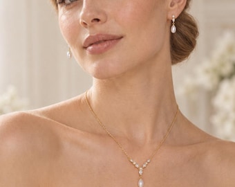 May include: A gold-toned necklace featuring a central teardrop-shaped white pearl pendant accented by small clear stones in a V-shaped arrangement, paired with matching gold-toned drop earrings, each featuring a single small clear stone and a pearl.