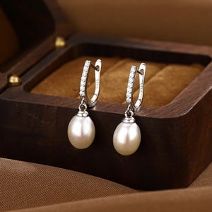 Puede incluir: Un par de pendientes de aro de plata con una sola perla blanca que cuelga de cada aro. Los aros est&aacute;n decorados con peque&ntilde;as piedras transparentes.
