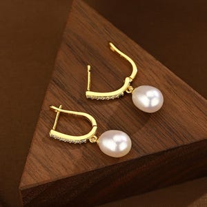 Puede incluir: Un par de pendientes de aro dorados con una sola perla blanca que cuelga de cada aro. Los aros est&aacute;n decorados con una fila de peque&ntilde;as piedras transparentes.