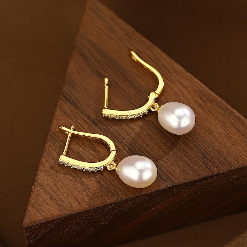 Puede incluir: Un par de pendientes de aro dorados con una sola perla blanca que cuelga de cada aro. Los aros est&aacute;n decorados con una fila de peque&ntilde;as piedras transparentes.