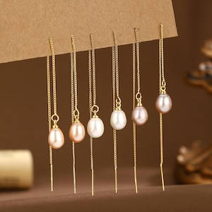 Puede incluir: Seis pares de pendientes de hilo dorados, cada uno con una perla de diferente color. Las perlas varían en color de blanco a rosa y melocotón. Los pendientes cuelgan de una superficie marrón.