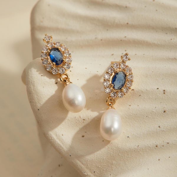 May include: A pair of gold-tone drop earrings featuring an oval blue center stone surrounded by a halo of clear crystals. Each earring is finished with a dangling white teardrop-shaped pearl suspended from the crystal-encrusted oval setting.