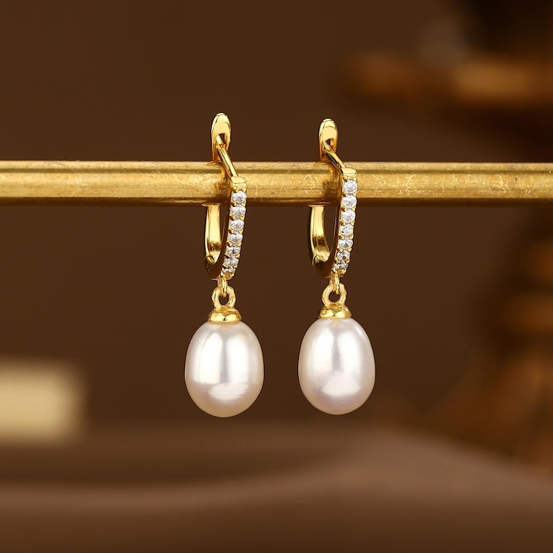 May include: A pair of gold hoop earrings with a single pearl drop on each earring. The hoops are adorned with small clear stones.