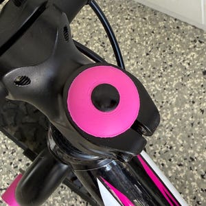 May include: Close-up of a bicycle handlebar with a black stem and bright pink accents. The handlebar grips and stem cap are pink, contrasting with the black frame. The bike frame has white and pink stripes.