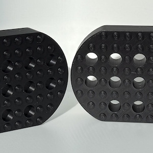 May include: Two black, rectangular, 3D-printed objects with rounded edges. Each has a textured surface with small, raised bumps and several circular holes. One object has a hexagonal hole. The objects are likely components or parts.