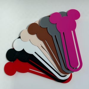 May include: A selection of colourful bookmarks in shades of red, black, white, beige, brown, grey, and pink. Each bookmark has a rounded top with two circular shapes, and a long, slender body. The bookmarks are arranged in a fan-like pattern.