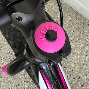 May include: Close-up of a bicycle handlebar with a pink accent. The handlebar is black with a pink circular accent that reads "KEIRA" in white letters. The bicycle frame has white and pink accents.