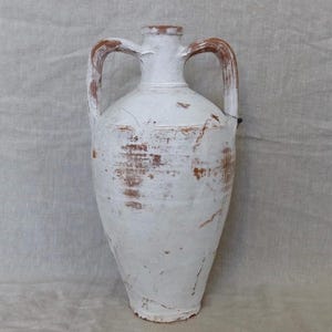 Puede incluir: Ánfora de cerámica blanca con dos asas, que muestra signos de desgaste y edad. La ánfora tiene un cuerpo redondeado y un cuello estrecho.