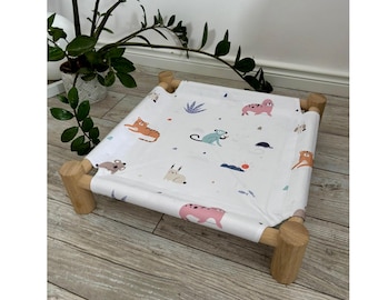 May include: A square pet bed featuring a light wood frame with four cylindrical legs and a white fabric sling. The fabric is printed with a colorful pattern of various animals including a tiger, monkey, seal, and rabbit, along with small sun icons.