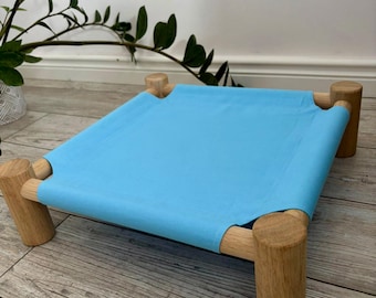 May include: A pet bed featuring a light blue fabric sleeping surface stretched across a frame made of four cylindrical light-toned wooden legs connected by horizontal wooden dowels, creating a low-profile elevated platform design.