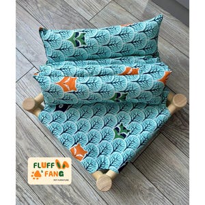 May include: A pet hammock with teal fabric patterned with trees and orange foxes. The hammock is supported by wooden dowels. The Fluff Fang logo is visible. The design is suitable for small pets.