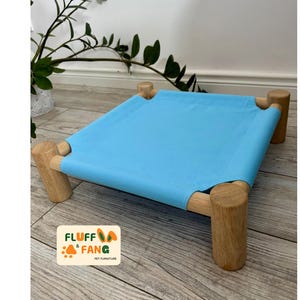 以下が含まれることがあります： 木製のフレームが付いた、水色のペット用ベッド。ベッドは長方形のデザインで、フレームに青い布が張られています。木製の脚は円筒形です。ペット用に設計されたベッドです。