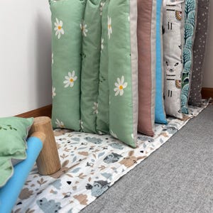 May include: A row of rectangular cushions standing upright on a patterned fabric base. The cushions feature various designs including white daisies on sage green, solid mauve, solid blue, llamas on a light background, and a grey star pattern.