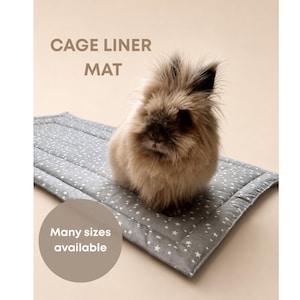 Pode incluir: Um tapete retangular cinzento para gaiola com um padrão de estrelas brancas. Um coelho castanho fofo está sentado no tapete. O texto CAGE LINER MAT e Many sizes available estão impressos na imagem. O tapete tem bordas costuradas e textura.