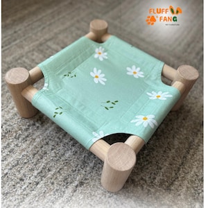 May include: A small pet bed with a light green fabric featuring white daisy and green leaf patterns. The bed is supported by four wooden cylindrical legs and a wooden frame. The brand name "Fluff Fang Pet Furniture" is visible.