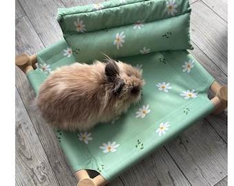 Peut inclure: Un petit lapin brun clair et duveteux repose sur un lit pour animal de couleur vert clair à motif de marguerites blanches. Le lit comprend un cadre en bois et un oreiller rectangulaire assorti, tous deux ornés du même imprimé floral.