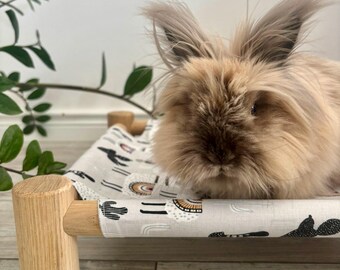 May include: A fluffy, light brown rabbit resting on a small, elevated pet bed. The bed features a light gray fabric cover with a repeating pattern of black cacti and colorful rainbows, supported by a light-colored wooden frame.