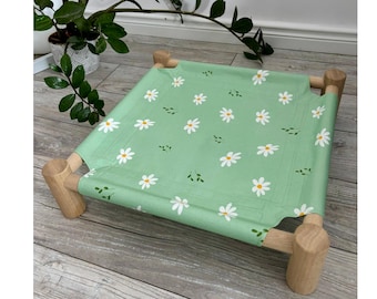 May include: A square pet bed featuring a light green fabric surface with a repeating pattern of white daisies and small green leaves, supported by a sturdy frame made of four cylindrical light-colored wooden legs at each corner.