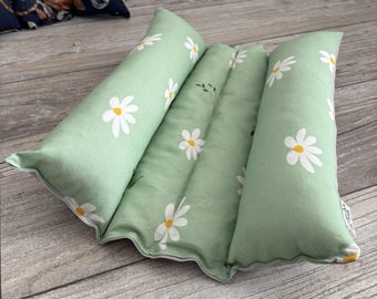 May include: A sage green fabric pillow featuring a repeating white daisy pattern with yellow centers. The pillow has a segmented, multi-chambered design with three distinct, elongated, stuffed sections and a small white brand tag on the side.