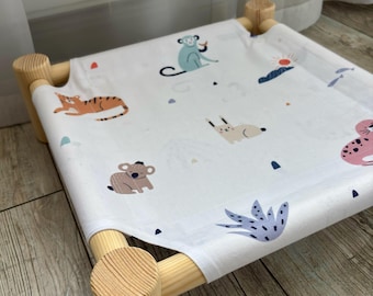 May include: A small pet bed with a light wood frame and a white fabric cover featuring a repeating pattern of colorful cartoon animals including a monkey, tiger, koala, and rabbit, along with small sun, plant, and mountain graphic elements.