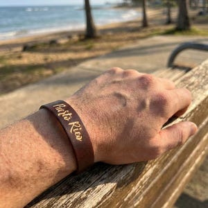 Puede incluir: Una pulsera de cuero marrón con las palabras "Puerto Rico" en letras doradas. La pulsera se lleva en la muñeca, con una playa y palmeras al fondo.