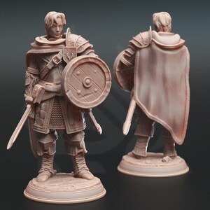 May include: A detailed figurine of a medieval warrior, clad in armor and holding a sword and shield. The figure is light brown, with intricate detailing on the armor, shield, and clothing. The warrior stands on a circular base, with a cape draped over the shoulders.