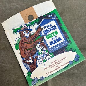 May include: Vintage Smokey Bear litter bag. The bag features a colorful illustration of Smokey Bear with the text "Keep America Green and Clean" and "Prevent Forest Fires". The bag is white with a brown top and a hole for hanging.