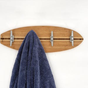 May include: A surfboard-shaped wooden wall hook with three metal cleats. The wood has a light brown finish with a dark brown stripe down the center. A blue towel hangs from the hooks.