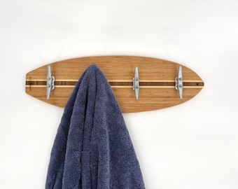 May include: A surfboard-shaped wooden wall hook with three metal cleats. The wood has a light brown finish with a dark brown stripe down the center. A blue towel hangs from the hooks.
