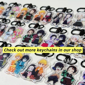 K&ouml;nnte beinhalten: Eine Sammlung von Anime-Charakter-Schl&uuml;sselanh&auml;ngern mit schwarzen Metallverschl&uuml;ssen. Die Schl&uuml;sselanh&auml;nger zeigen farbenfrohe Illustrationen verschiedener Charaktere. Text auf einem gelben Banner: "Check out more keychains in our shop."