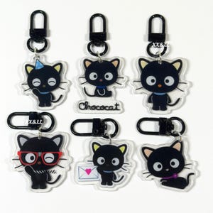 May include: A collection of black cat-shaped keychains with various accessories and expressions. Each keychain features a black cat design with yellow accents, attached to a black metal clasp. Some cats wear glasses, a party hat, or hold an envelope.