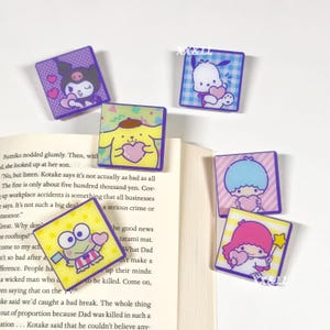 May include: A collection of six square acrylic charms featuring various cartoon characters. Each charm has a different character design, including a purple Kuromi, a white Pochacco, a yellow Pompompurin, a green Keroppi, and two Little Twin Stars. The charms are on a book.