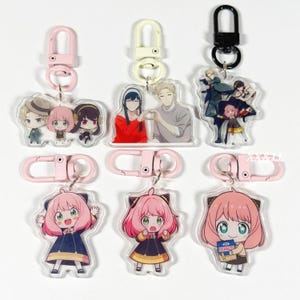 May include: Collection of acrylic keychains featuring anime characters. The keychains have pink, white, and black clasps. The characters have pink hair, black hair, and wear various outfits. One keychain shows a character holding a snack.