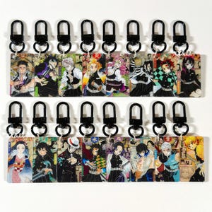May include: A collection of sixteen rectangular acrylic keychains featuring various anime characters from Demon Slayer, each attached to a black swivel lobster clasp. The designs display colorful illustrations of characters in uniform with swords.