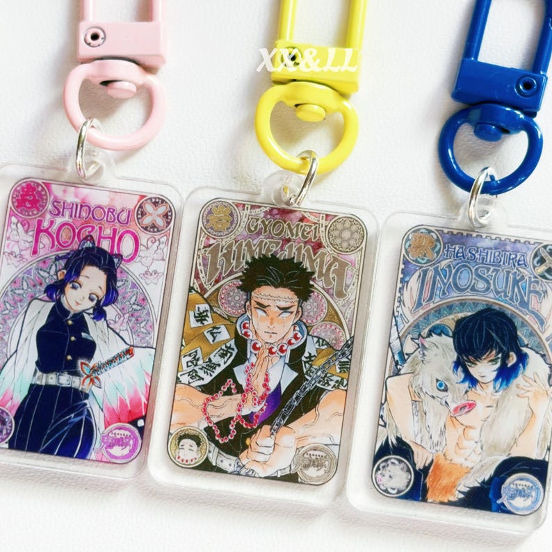 K&ouml;nnte beinhalten: Drei Schl&uuml;sselanh&auml;nger aus Acryl mit farbenfrohen Illustrationen von Anime-Charakteren. Jeder Schl&uuml;sselanh&auml;nger hat ein anderes Charakterdesign und einen farblich passenden Verschluss in Rosa, Gelb und Blau. Die Schl&uuml;sselanh&auml;nger zeigen japanischen Text und dekorative Bord&uuml;ren.