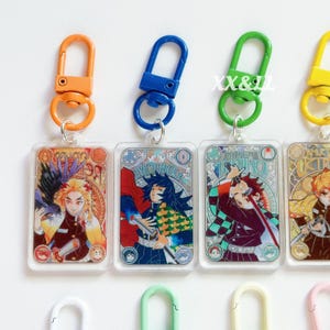 K&ouml;nnte beinhalten: Schl&uuml;sselanh&auml;nger mit Anime-Charakter-Illustrationen. Jeder Schl&uuml;sselanh&auml;nger hat einen rechteckigen Acryl-Anh&auml;nger mit einem Charakterdesign und einen Metallverschluss in verschiedenen Farben, darunter Orange, Blau und Gr&uuml;n. Die Schl&uuml;sselanh&auml;nger sind an einem silbernen Ring befestigt.