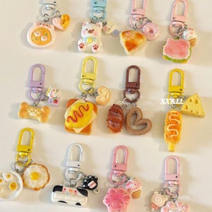 May include: A collection of colorful keychains with various food and animal-themed charms. The charms include a bear, a cow, a paw, a slice of cheese, and miniature pastries. Each keychain has a metal clasp.