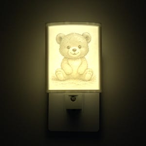 Peut inclure: Veilleuse émettant une lumière douce et chaleureuse. Elle présente une forme rectangulaire avec une illustration détaillée d'un ours en peluche. L'ours est assis et souriant, de couleur beige clair. La veilleuse est branchée sur une prise murale.