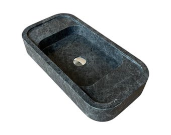 May include: Dark grey rectangular bathroom sink with a rounded rim and a central drain. The sink has a textured surface and a built-in shelf around the basin. The sink is made of stone.