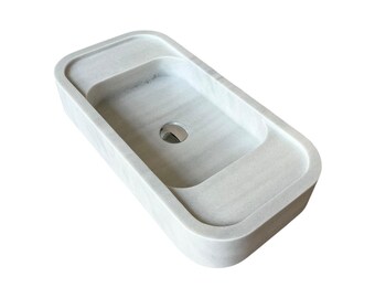 May include: A rectangular, light gray marble sink with a rounded rim. The sink has a central drain and a raised area on one side. The overall design is minimalist and modern, suitable for a bathroom.