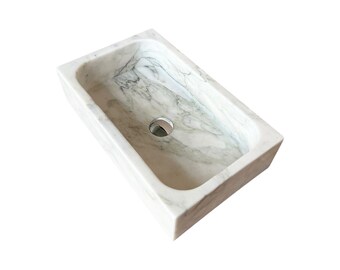 May include: Rectangular marble sink with a white and grey veined pattern. The sink has a drain hole in the centre and is made of a solid block of stone. The design is modern and minimalist, suitable for a bathroom.