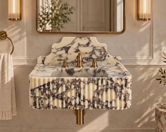 Puede incluir: Un lavabo de pared hecho de mármol blanco con vetas moradas, que presenta un diseño frontal estriado y un salpicadero festoneado. El lavabo incluye un grifo central de latón con dos manijas laterales a juego y un tubo de drenaje de latón.
