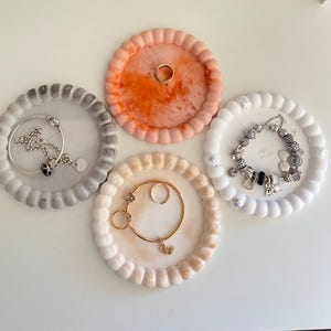 May include: Four decorative jewellery trays with beaded edges. One tray is grey, holding a silver necklace. Another is peach, with a gold ring. A third is white, with a charm bracelet. The last is beige, with gold rings.