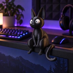 May include: A black cat figurine with large white eyes and pointed ears, designed to sit on the edge of a computer monitor. The cat has a smooth, matte finish, a slender body, and a long tail that curves downward over the edge of the screen.
