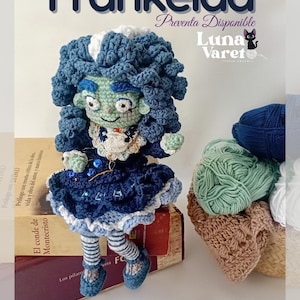 Puede incluir: Muñeca de ganchillo hecha a mano llamada "Frankelda" con cabello azul y un vestido azul. La muñeca está sentada sobre una pila de libros. La imagen incluye el texto "Frankelda", "Luna Varet" y "Preventa Disponible".