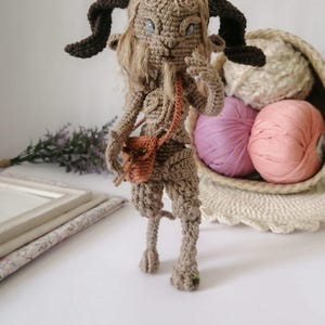 May include: A crocheted figure of a mythical creature with brown fur, dark horns, and a small orange bag. The figure has long hair and blue eyes. The background includes a basket with yarn and a decorative frame.