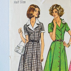 May include: A vintage sewing pattern envelope labeled 6157 Simplicity, featuring illustrations of two dresses. One is a black and white checkered dress with a white collar, and the other is a solid green dress with buttons. Text includes Size 18 1/2, Bust 41, and Look Slimmer pattern.